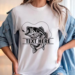 May include: White t-shirt featuring a black and white graphic of a largemouth bass with an open mouth and a fishing hook. The text "TEXT HERE" is displayed in a horizontal black bar below the fish.