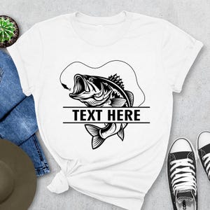 May include: White t-shirt featuring a black and white graphic of a fish with an open mouth, a fishing hook, and the text "TEXT HERE" in a bold font. The design is centered on the shirt.