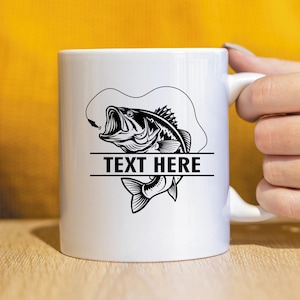May include: White ceramic mug featuring a black and white illustration of a largemouth bass with a fishing lure. The text "TEXT HERE" is displayed in a horizontal bar below the fish. The mug has a white handle.