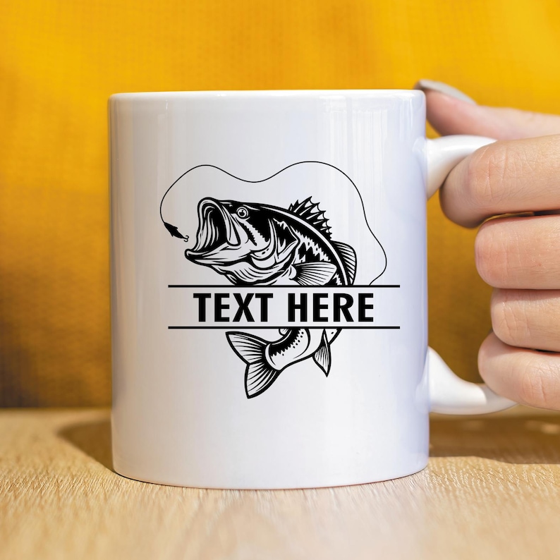 May include: White ceramic mug featuring a black and white illustration of a largemouth bass with a fishing lure. The text "TEXT HERE" is displayed in a horizontal bar below the fish. The mug has a white handle.