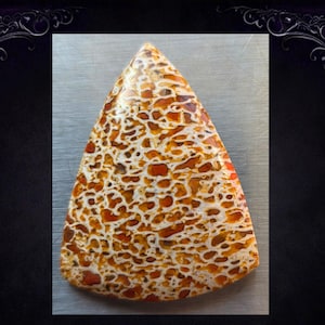 Puede incluir: Una gema triangular pulida con un patrón único. La piedra presenta una red de vetas blancas y marrón-anaranjadas, creando un diseño orgánico llamativo. La gema se encuentra sobre una superficie metálica, resaltando su belleza natural.