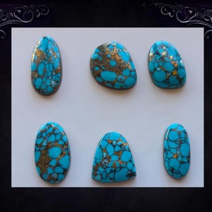 May include: Six turquoise and gold-colored gemstone cabochons in various shapes, including oval and teardrop. The stones have a vibrant blue hue with intricate gold veining patterns. The gemstones are displayed on a white background.