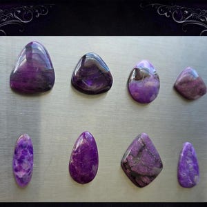 May include: Eight polished purple gemstones of varying shapes, including oval, teardrop, and diamond, are arranged on a silver surface. The stones display a range of purple hues, from deep violet to lighter lavender, with visible patterns and textures.