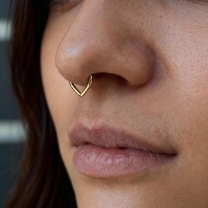 Puede incluir: Primer plano de una nariz con un aro de septum dorado. El aro tiene forma de V, con la punta hacia abajo. El tono de piel es claro y los labios son visibles.