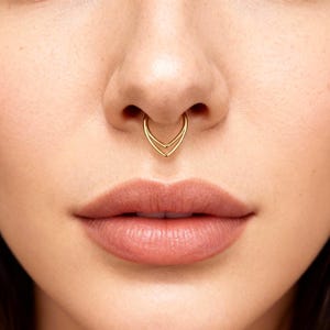 Könnte beinhalten: Ein goldfarbener Septum-Piercing mit doppeltem Chevron-Design. Der Schmuck wird an der Nase einer Person getragen, wobei Lippen und Haut sichtbar sind. Das Piercing ist ein stilvolles Accessoire.