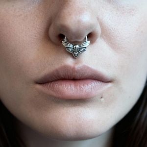 May include: A silver septum ring with a moth design. The piercing is positioned on the nose, with the moth's wings displayed. The image is a close-up, highlighting the jewellery and the lower face.