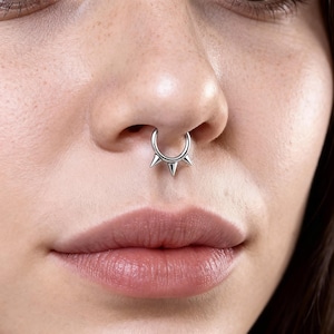 Op de afbeelding: Een zilveren septumring met drie conische spikes. De sieraden worden gedragen in een doorboorde neus, wat een moderne en edgy uitstraling geeft. De ring is een opvallend accessoire dat een vleugje punk of alternatieve stijl toevoegt.