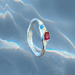 May include: A silver ring with a unique, organic design. It features a rectangular red gemstone and a small, round blue gemstone. The ring is set against a blurred, light blue background.