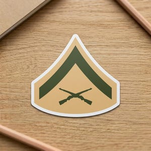 May include: A tan and green sticker featuring a military rank insignia. The design includes two crossed rifles beneath a green chevron on a tan background. The sticker is on a wooden surface.