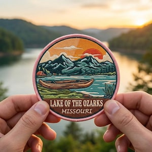 May include: Round embroidered patch featuring a scenic landscape of the Lake of the Ozarks in Missouri. The design includes a lake, mountains, a canoe, and a sunset. The patch has a pink border and the text "Lake of the Ozarks Missouri".