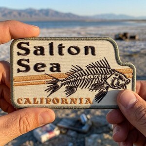 May include: Embroidered patch featuring the words "Salton Sea California" above a detailed illustration of a fish skeleton. The patch has a beige background with brown and green accents, and is held up by a person's hands.