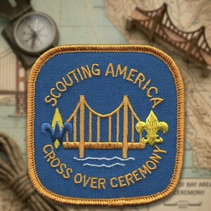 May include: A blue embroidered patch with the words "Scouting America Cross Over Ceremony" in gold lettering. The patch features a gold bridge design and a gold fleur-de-lis symbol. The patch has a gold border.