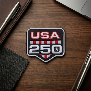 May include: A black, white, and red patch with the text "USA 250" and a star. The patch has a shield shape and is on a wooden surface. The patch is surrounded by a pen and a notebook.