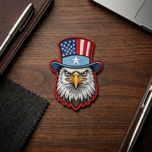 May include: Embroidered patch featuring an eagle's head wearing a patriotic top hat. The hat displays the American flag design with stars and stripes. The eagle has a stern expression, with a yellow beak and white and gray feathers, outlined in red.