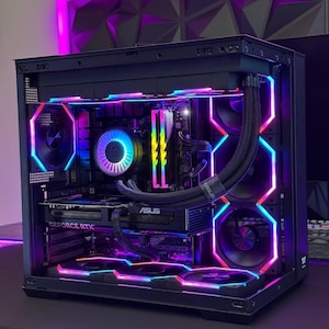May include: A black computer case with internal components illuminated by vibrant RGB lighting. The case features a GeForce RTX graphics card and an ASUS component. The lighting includes shades of blue, pink, and purple, creating a visually striking display.