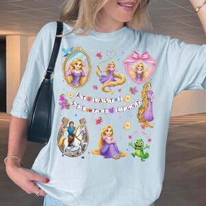 May include: A light blue t-shirt featuring a graphic print of Rapunzel, a horse, and a chameleon. The design includes various illustrations of the character, floral accents, and the text AT LAST I SEE THE LIGHT in circular bubbles across the center.