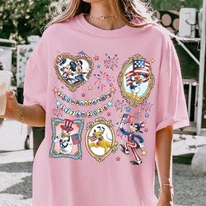 May include: Pink t-shirt featuring cartoon characters in patriotic attire. The shirt has several framed images of characters like Mickey Mouse, Donald Duck, and Pluto, celebrating the 250th anniversary of 1776-2026. Fireworks and stars are also included in the design.