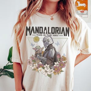 以下が含まれることがあります： クリーム色のTシャツで、マンダロリアンとグローグーが色とりどりの花、蝶、惑星に囲まれたグラフィックプリントが施されています。中央のキャラクターイラストの上部には、黒い文字で「MANDALORIAN THIS IS THE WAY」とプリントされています。