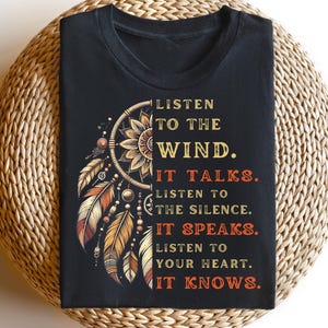 Könnte beinhalten: Schwarzes T-Shirt mit einem Traumfänger-Design und orangefarbenen, braunen und gelben Federn. Der Text auf dem Shirt lautet: "Höre auf den Wind. Er spricht. Höre auf die Stille. Sie spricht. Höre auf dein Herz. Es weiß."