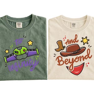 May include: Two t-shirts, one green and one cream-colored, with cartoon graphics. The green shirt has a Buzz Lightyear-themed design with the text "To Infinity". The cream shirt features a cowboy hat, boot, and the words "And Beyond".