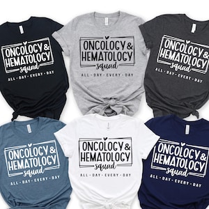 Może przedstawiać: Sześć koszulek w różnych kolorach: czarnym, szarym, antracytowym, niebieskim, białym i granatowym. Każda koszulka ma napis "ONCOLOGY & HEMATOLOGY squad" w białym prostokącie, z napisem "ALL DAY EVERY DAY" poniżej.