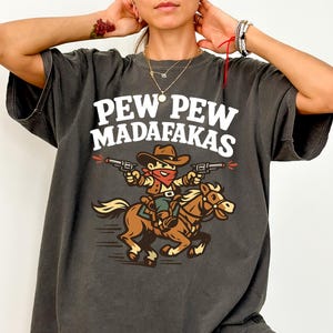 May include: Dark grey t-shirt featuring a cartoon cowboy riding a horse and shooting guns. The shirt has the text "PEW PEW MADAF*KAS" in white block letters above the graphic. The cowboy is wearing a brown hat and red bandana.