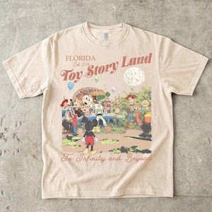 May include: A beige short-sleeve t-shirt featuring a graphic print with the text "FLORIDA Est. 2018 Toy Story Land" and "To Infinity and Beyond". The illustration depicts Mickey Mouse, Woody, Buzz Lightyear, and Slinky Dog in a park setting.