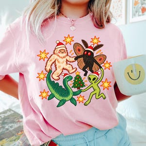 May include: A light pink t-shirt featuring a graphic of a white yeti, a brown moth, a green dinosaur, and a green alien, all wearing Santa hats and dancing around a small Christmas tree, surrounded by yellow star shapes.