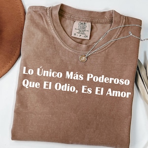 May include: A brown Comfort Colors t-shirt with the text "Lo Único Más Poderoso Que El Odio, Es El Amor" in white. A silver necklace with a small pendant rests on the shirt. A brown hat and dried leaves are also visible.
