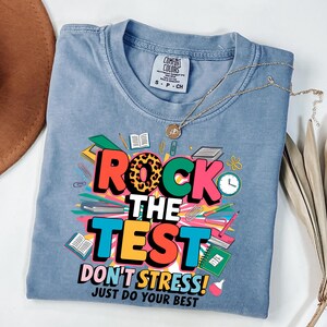 Op de afbeelding: Een lichtblauw t-shirt met de tekst "ROCK THE TEST" in een kleurrijk ontwerp. Het shirt bevat ook de zinnen "DON'T STRESS!" en "JUST DO YOUR BEST". Een gouden ketting is over het shirt gedrapeerd.