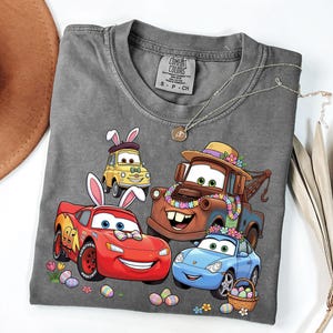 Può includere: Una maglietta grigia Comfort Colors con una grafica colorata di auto dei cartoni animati decorate per Pasqua. Le auto includono Saetta McQueen, Cricchetto e altri, con uova di Pasqua e fiori. Sono visibili anche una collana e un cappello.