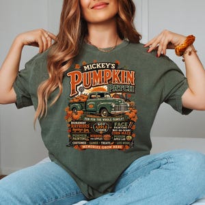 Könnte beinhalten: Ein dunkelolivgrünes T-Shirt mit einem Grafikdruck eines Oldtimer-Lastwagens, der mit Kürbissen beladen ist. Der Text lautet Mickey's Pumpkin Patch, Fun For The Whole Family, und listet Aktivitäten wie Hayrides, Corn Maze und Memories Grow Here auf.