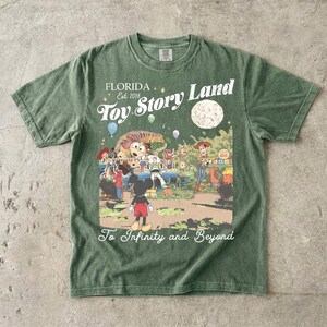 May include: A sage green short-sleeve t-shirt featuring a graphic print with the text Florida Est 2018 Toy Story Land and To Infinity and Beyond. The illustration depicts Mickey Mouse, Woody, Buzz Lightyear, and Slinky Dog in a park setting.
