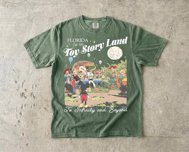 May include: A sage green short-sleeve t-shirt featuring a graphic print with the text Florida Est 2018 Toy Story Land and To Infinity and Beyond. The illustration depicts Mickey Mouse, Woody, Buzz Lightyear, and Slinky Dog in a park setting.