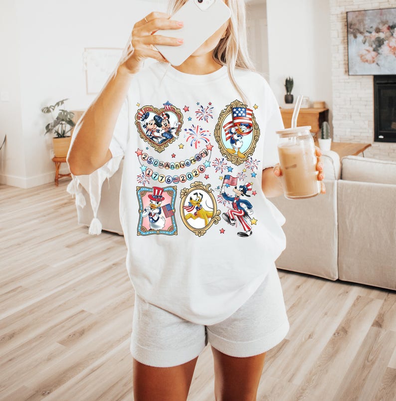 May include: White t-shirt featuring patriotic cartoon characters in ornate frames. The design includes Mickey Mouse, Donald Duck, and other characters celebrating the 250th anniversary of 1776-2026. The shirt has a relaxed fit.