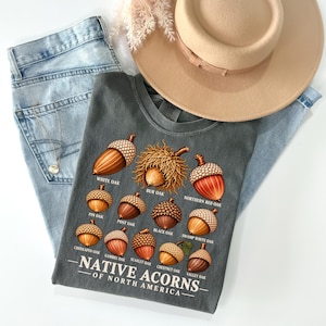 May include: A charcoal gray t-shirt featuring a graphic of various acorns labeled with their names, including White Oak, Bur Oak, and Northern Red Oak, with the text Native Acorns of North America, paired with light blue jeans and a tan hat.
