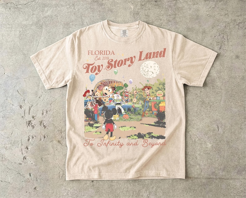 May include: A beige short-sleeve t-shirt featuring a graphic print with the text "FLORIDA Est. 2018 Toy Story Land" and "To Infinity and Beyond". The illustration depicts Mickey Mouse, Woody, Buzz Lightyear, and Slinky Dog in a park setting.