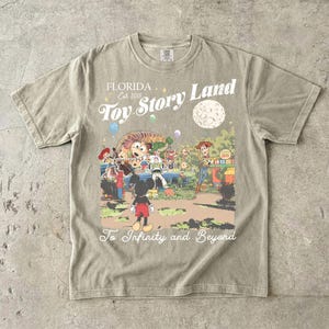 May include: A beige short-sleeve t-shirt featuring a graphic print of Toy Story Land with characters including Woody, Buzz Lightyear, and Mickey Mouse. The text reads Florida Est 2018, Toy Story Land, and To Infinity and Beyond.