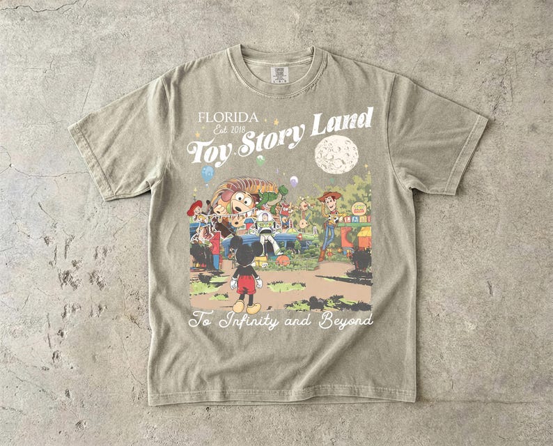 May include: A beige short-sleeve t-shirt featuring a graphic print of Toy Story Land with characters including Woody, Buzz Lightyear, and Mickey Mouse. The text reads Florida Est 2018, Toy Story Land, and To Infinity and Beyond.