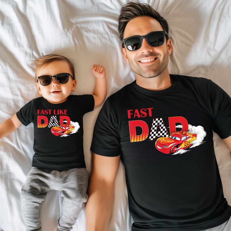 May include: Matching black t-shirts with the text "FAST DAD" and a cartoon race car graphic. One shirt is for an adult, the other for a child. Both are wearing sunglasses and lying on a white bedsheet.