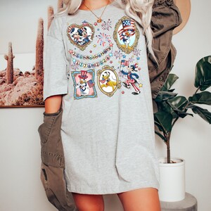 May include: A light gray t-shirt featuring framed cartoon characters celebrating the 250th anniversary of 1776-2026. The frames include Mickey and Minnie Mouse, Donald Duck, and Pluto, with patriotic themes and fireworks.