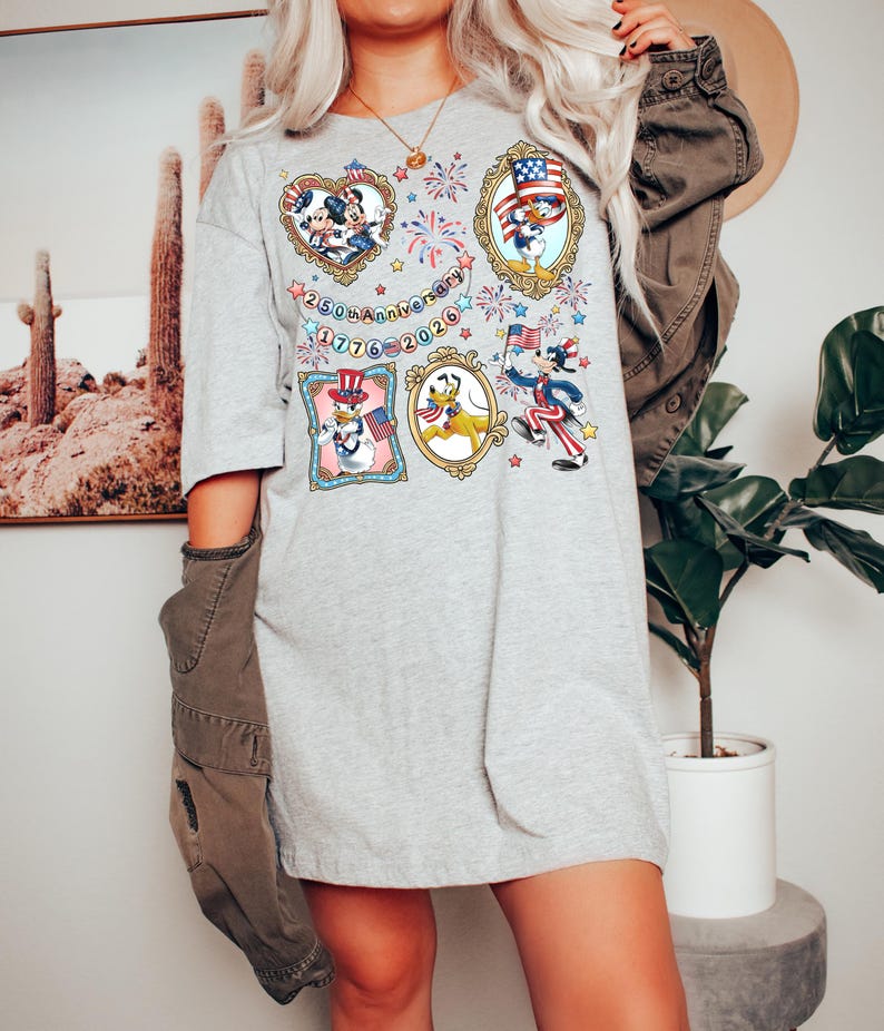 May include: A light gray t-shirt featuring framed cartoon characters celebrating the 250th anniversary of 1776-2026. The frames include Mickey and Minnie Mouse, Donald Duck, and Pluto, with patriotic themes and fireworks.