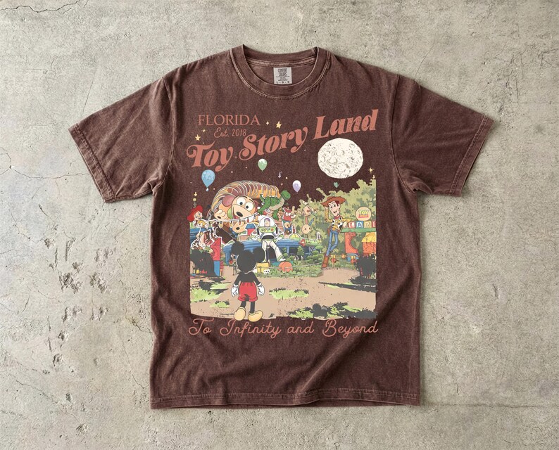 May include: A brown short-sleeve t-shirt featuring a graphic print with the text "FLORIDA Est. 2018 Toy Story Land" and "To Infinity and Beyond". The illustration depicts Mickey Mouse, Woody, Buzz Lightyear, and Slinky Dog under a large moon.