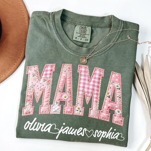 Puede incluir: Camiseta verde oliva Comfort Colors con la palabra "MAMA" en letras de patchwork rosa. Las letras están decoradas con estampados florales y de cuadros. Debajo de la palabra "MAMA" están los nombres "olivia james sophia" en escritura blanca.