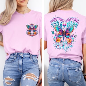 May include: A light pink t-shirt featuring a colorful butterfly design on the front pocket and back. The back of the shirt has the text "Everything Happens For A Reason, Just Believe" in a whimsical font. The butterfly has blue, orange, and yellow accents.