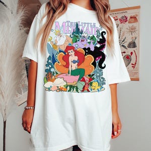May include: White t-shirt featuring a colorful graphic of characters from "The Little Mermaid." The design includes Ariel, King Triton, Ursula, and other characters. The title "The Little Mermaid" is printed in a stylized font.