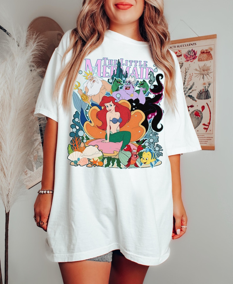 May include: White t-shirt featuring a colorful graphic of characters from "The Little Mermaid." The design includes Ariel, King Triton, Ursula, and other characters. The title "The Little Mermaid" is printed in a stylized font.