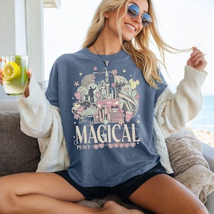 May include: A blue t-shirt with a colorful graphic design featuring a castle, flowers, and the text "The Most Magical Place". The design includes pink, yellow, and white elements. The shirt is worn by a person.