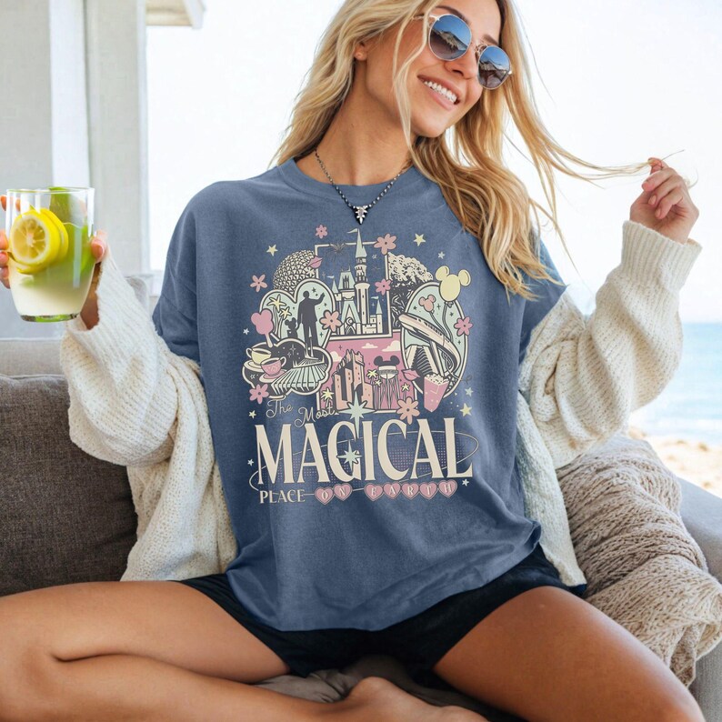 May include: A blue t-shirt with a colorful graphic design featuring a castle, flowers, and the text "The Most Magical Place". The design includes pink, yellow, and white elements. The shirt is worn by a person.