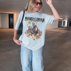 以下が含まれることがあります： ライトブルーのTシャツ。マンダロリアンとグローグーのグラフィックプリントが施され、周囲には色とりどりの花と蝶が描かれています。キャラクターの上部には、細い金の三角形の中に「MANDALORIAN」と「THIS IS THE WAY」という黒い文字がプリントされています。